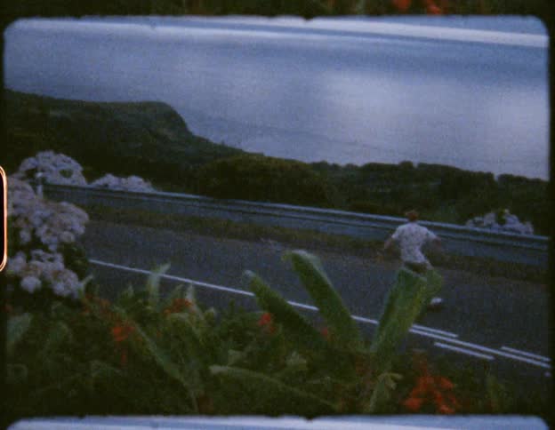 A man rides a skateboard back and forth down a narrow road just over the ocean