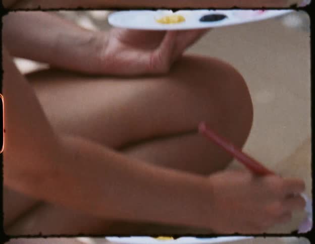 Woman sitting down holding a tray with paint in one hand while she paints on a canvas with a paintbrush with the other hand