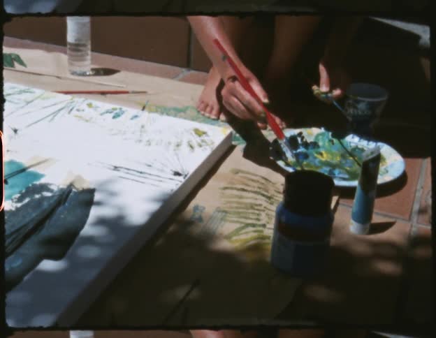 Person adds paint to narrow tool near large canvas set on floor