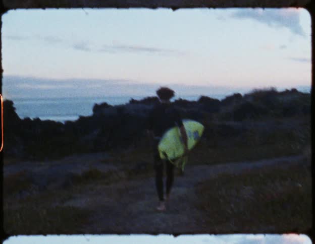 A man near the coast walking on a path holding a surfboard with the ocean the sky and clouds in the background