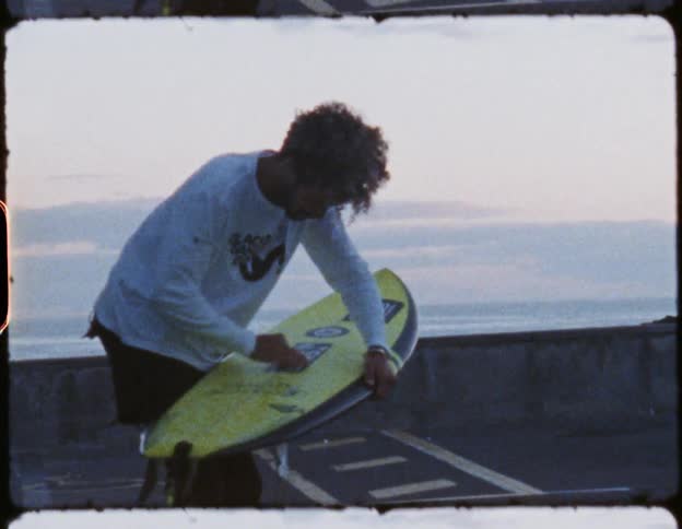 A surfer stands in a parking lot while thoroughly wiping down his surfboard with wax