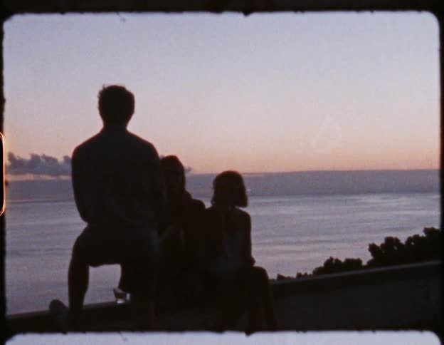 Three people sit on low wall overlooking ocean stretching to pink tinted horizon