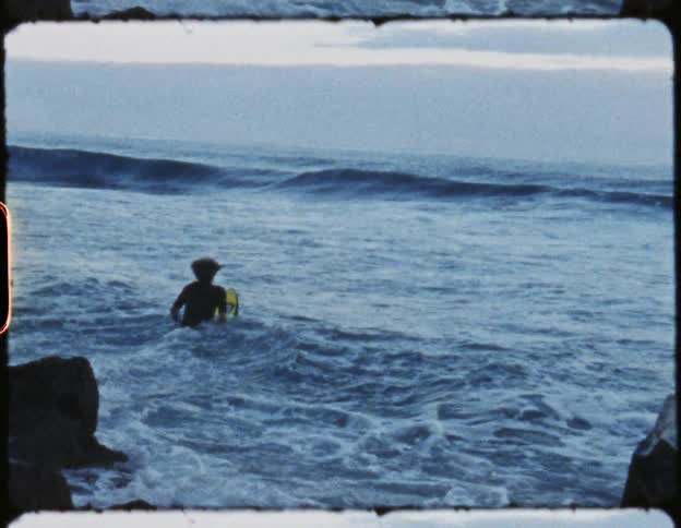 A surfer leaps with their surfboard off a rock formation into the ocean and swims toward the incoming waves