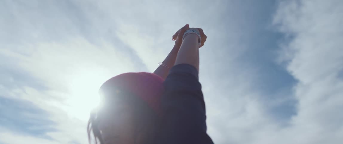 A woman with a hat and a smart watch standing and stretching with the sky and clouds in the background