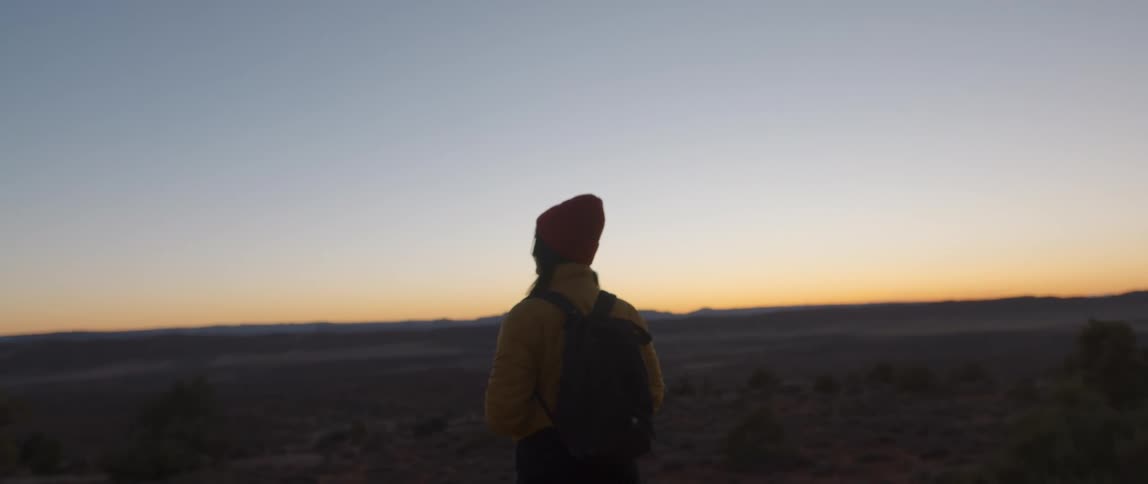 A woman hiking through a shrubland at sunrise pauses to gaze into the distance