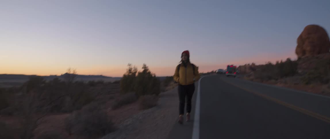 A woman with a hat a backpack and jacket walking in the side of a desert road with the sky and clouds in the background