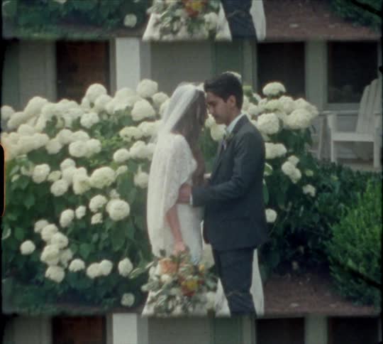 A man and a woman wearing bride and groom attire kiss one another in front of a house