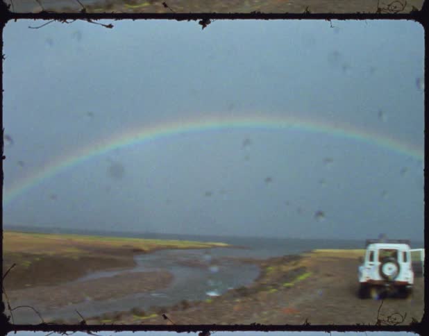 A rainbow is against a blue sky and landscape with a white jeep parked next to a creek and brown grass