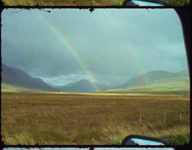 A double rainbow is showing in the middle of a grass field to the left of a vehicle