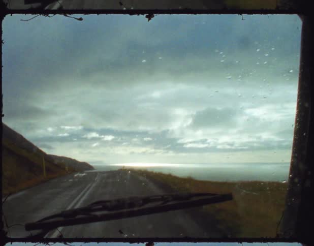 A vehicle travels along a coastal road that overlooks the ocean on a rainy day