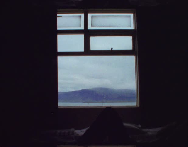 A woman lies in bed with rolling hills and a waterway outside the window of her home