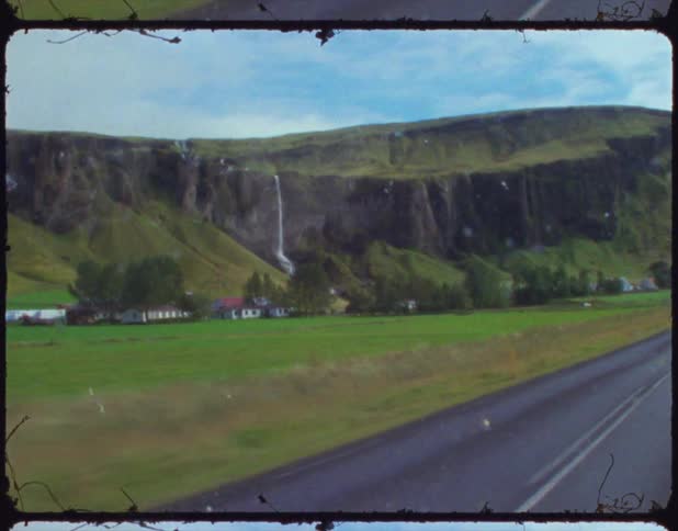 A shot from inside a moving car on a paved road with a hillside and a waterfall to the left of the road