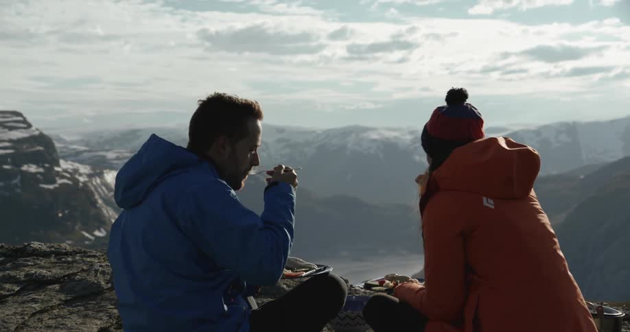 A man and woman eat while sitting near the edge of a mountain in the winter
