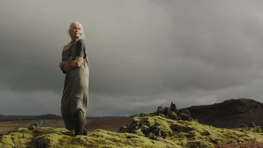 An elderly woman hugs her body as she stands in the wind on a moss covered hill