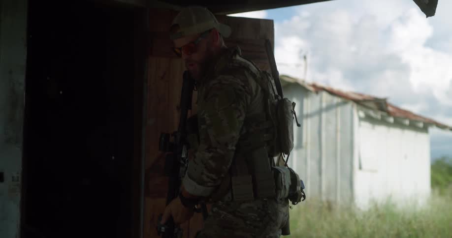 A military personnel is inside a barn while another man walks outdoors between the barn and a white shed during the day