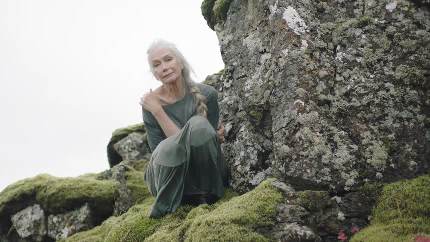 An elderly woman knees on the edge of a mossy cliff