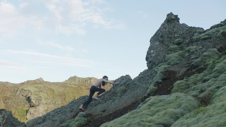 Shirtless long haired young man climbing barefoot over the moss covered rocks of a hill in the highlands