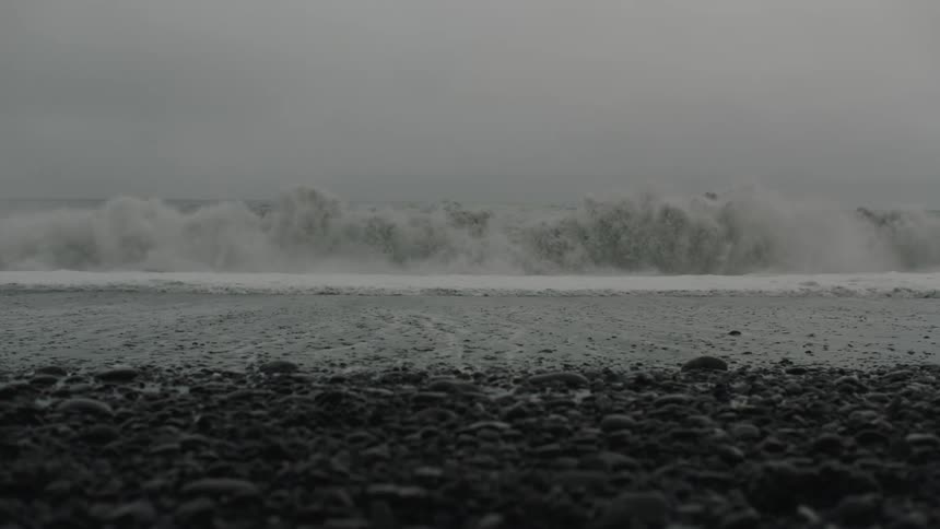 A sizable wave crashes down upon a beach lined with smooth stones