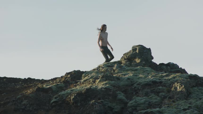 Young shirtless long haired man running on the black rocks on a mountain