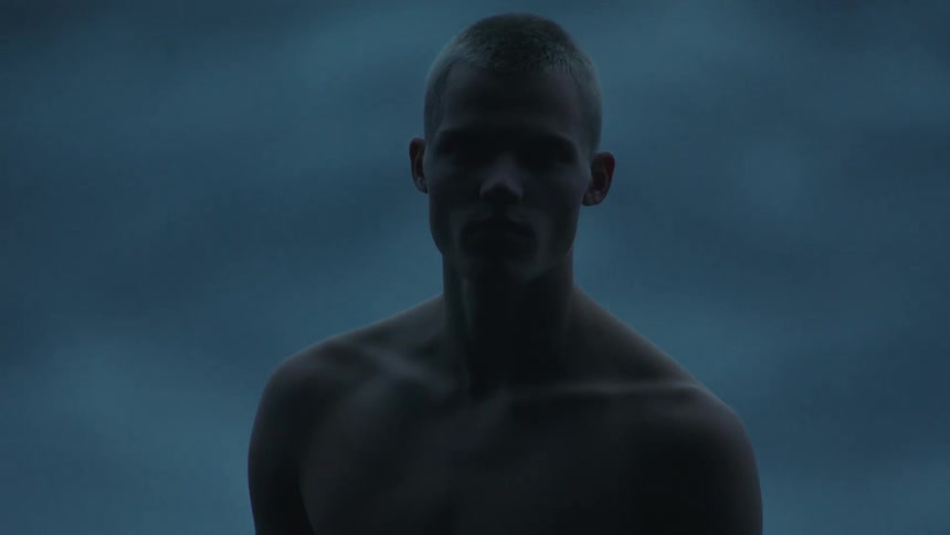A serious shirtless young man stares into the camera while cloaked in shadow