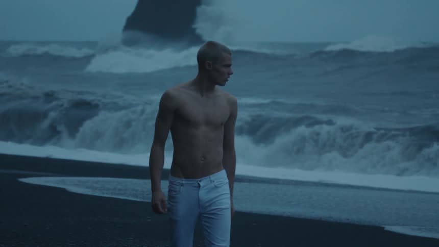 A young shirtless man walks along a wavy beach next to the ocean