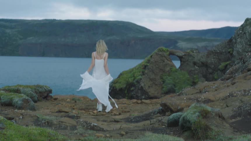 Young blonde caucasian woman in a white dress and shawl with arms outstretched walking down a hill to a lake