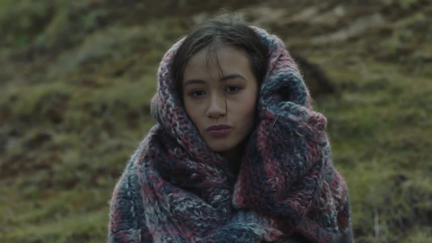 Young woman looking at camera covers herself with a colorful blanket near the hills on the highlands