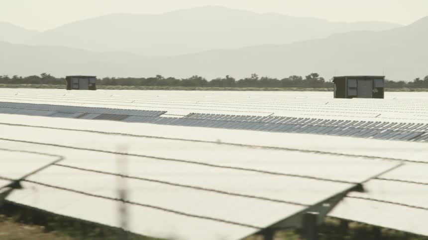 Rows of solar panels sit outside on a field with hills in the background