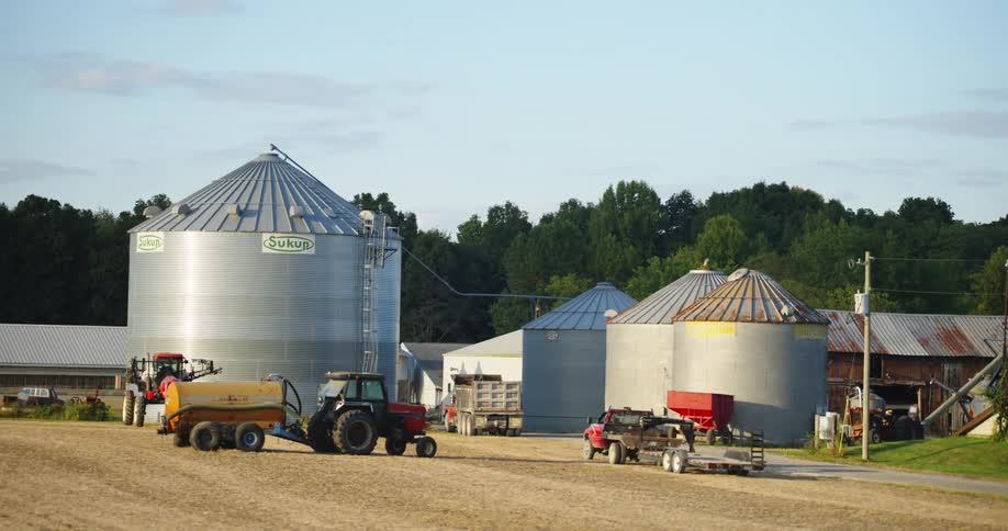 Trucks and tractors on a farm by silos on a sunny day with buildings and trees in the background