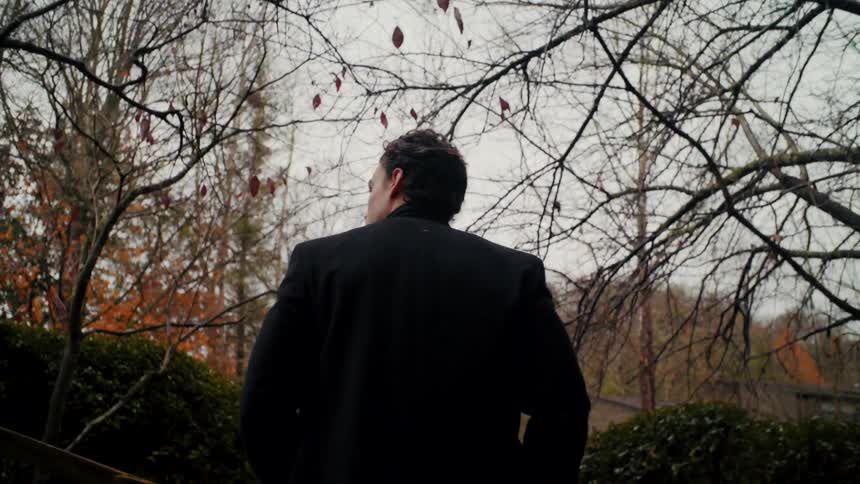 A man with a jacket walking in a pathway surrounded by trees with the sky and clouds in the background