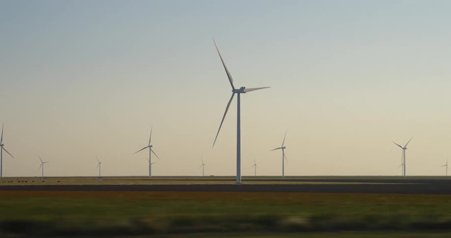 A road with vehicles next to a grassy plain with a wind turbine farm with the sky and clouds in the background