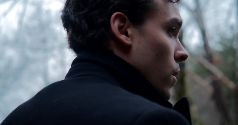 A man adjusts the collar of his coat while standing in a woodland during the day