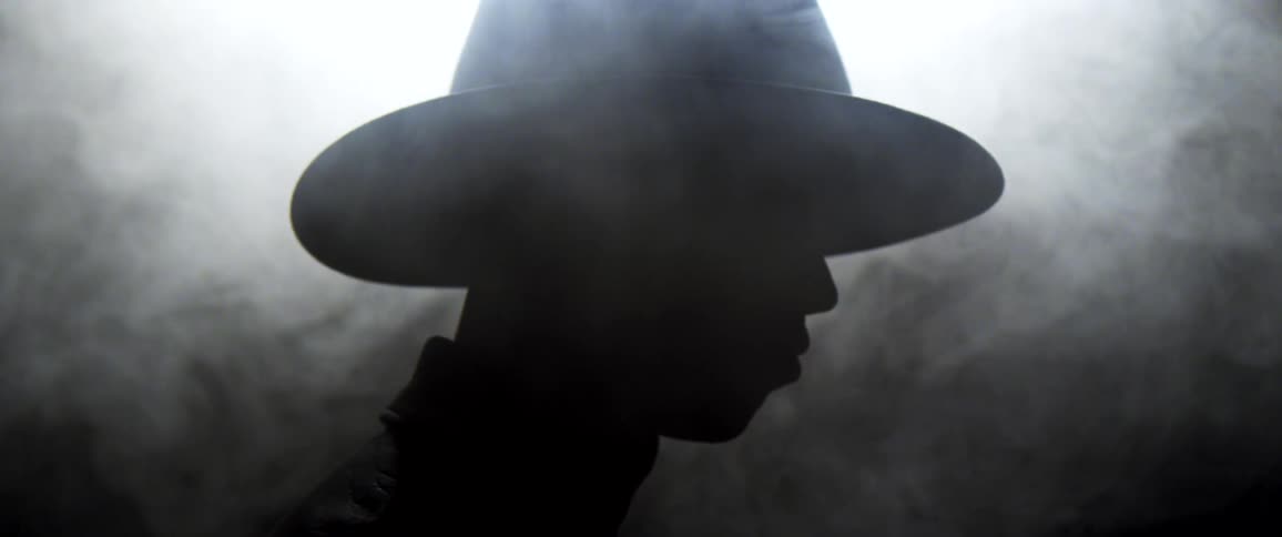 The silhouette of a person wearing a wide brimmed hat cloaked in swirling smoke under overhead lighting in a dark room