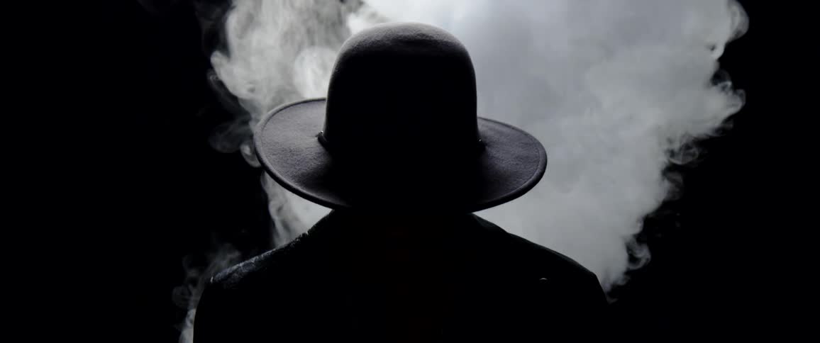 Silhouette of a person wearing a wide brim hat in front of a column of white smoke in a dark room