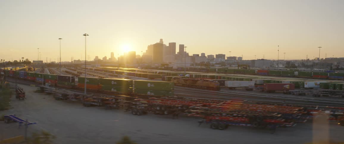 Train tracks with parked cargo cars in a lot with a city in background with the sun setting