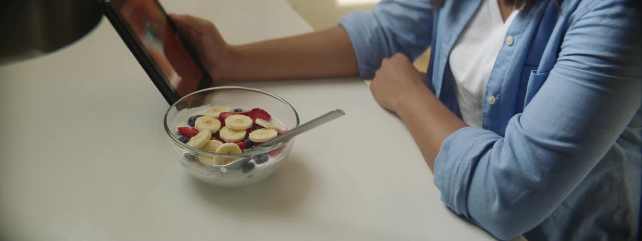 A woman sitting by a table with a bowl of milk and fruits looking at a smart phone
