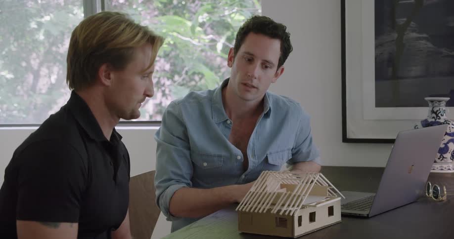 A man talks to his client as they sit at a table with a model of a house and a laptop
