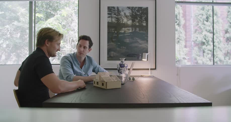 Two men sit together at a table with a model building structure and shake hands