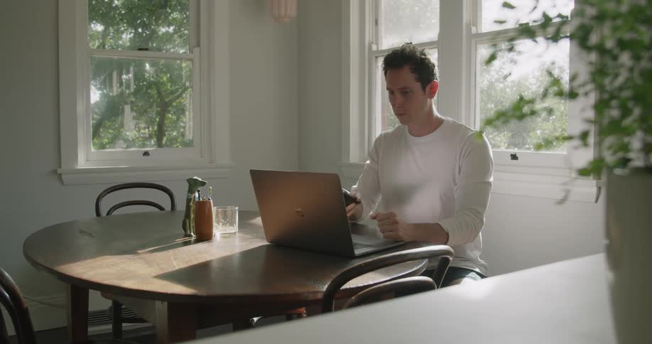 A man sits at a table at home talking on phone and then working on a laptop in front of him