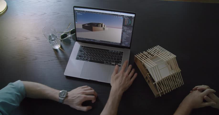 Two coworkers sitting at a table compare a physical model of a house to a rendition on a laptop