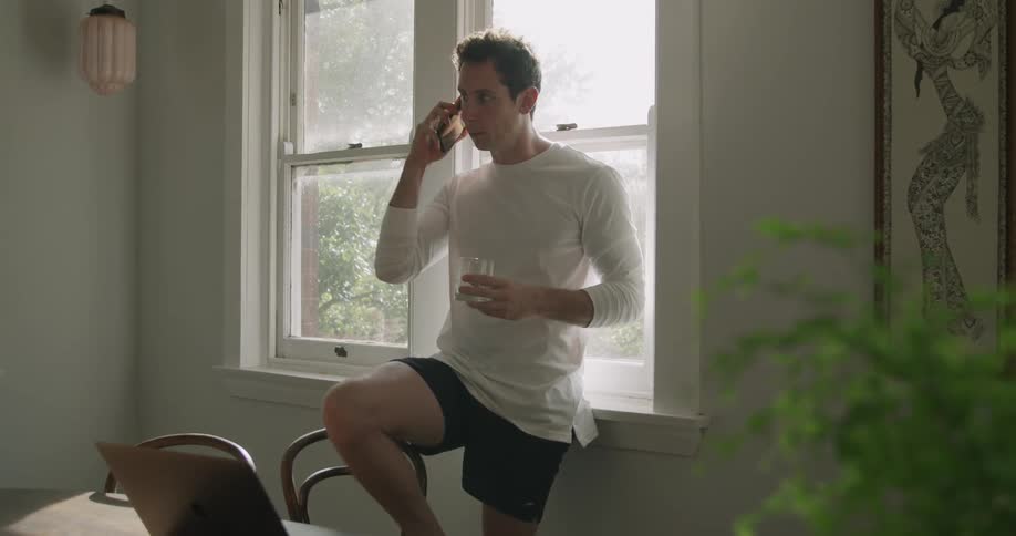 Young man reclined on a window in a room answers a call on a smartphone while working on a laptop