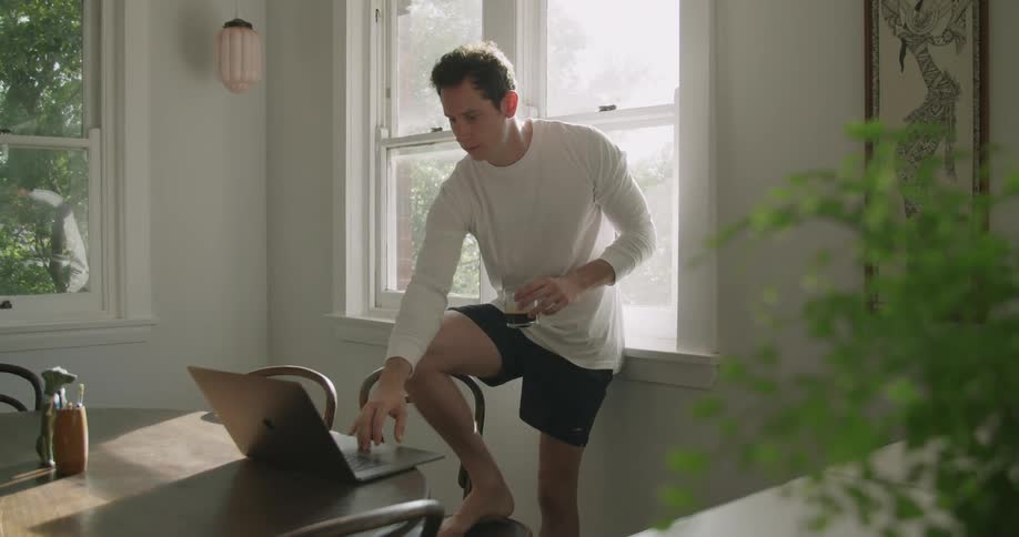 A man standing in a house with his foot on a chair drinks from a glass before leaning forward to type on a laptop