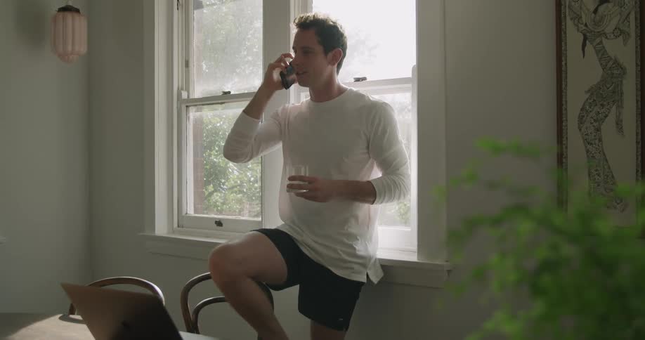 A man standing with one foot atop a chair while leaning against a window talks on a smartphone