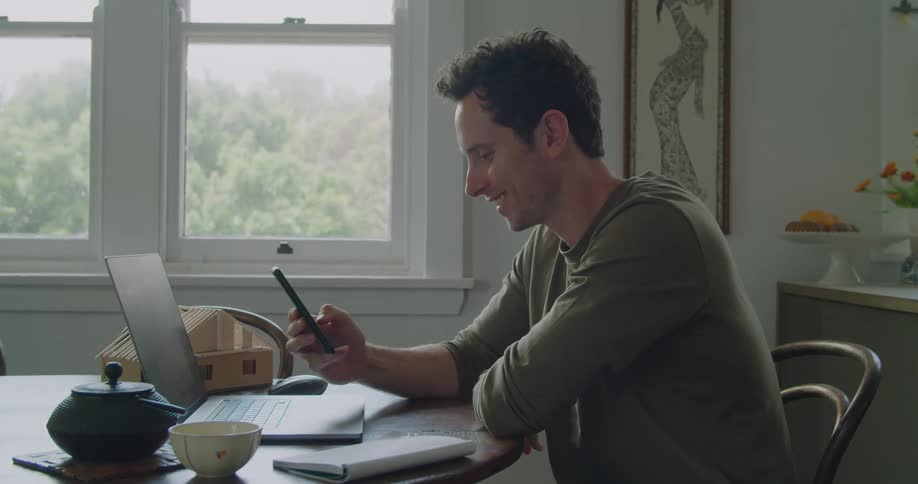 A man laughs while scrolling through his smartphone while sitting at his dining room table