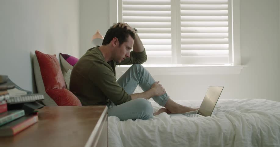 Young man sitting on the bed at home uses a laptop