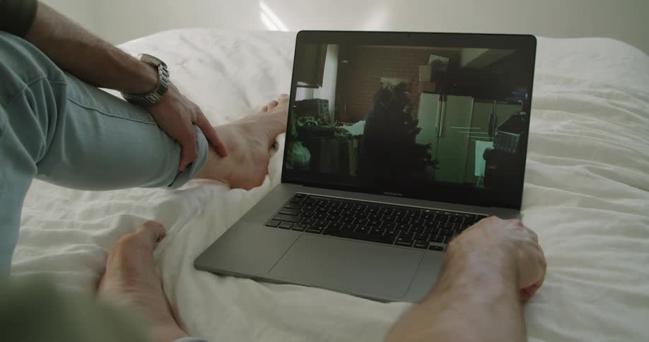 A person with a wristwatch sitting on a bed looking at a laptop