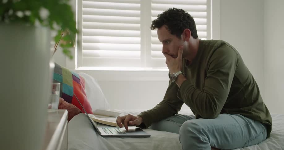 A man sits on his bed and types on his laptop