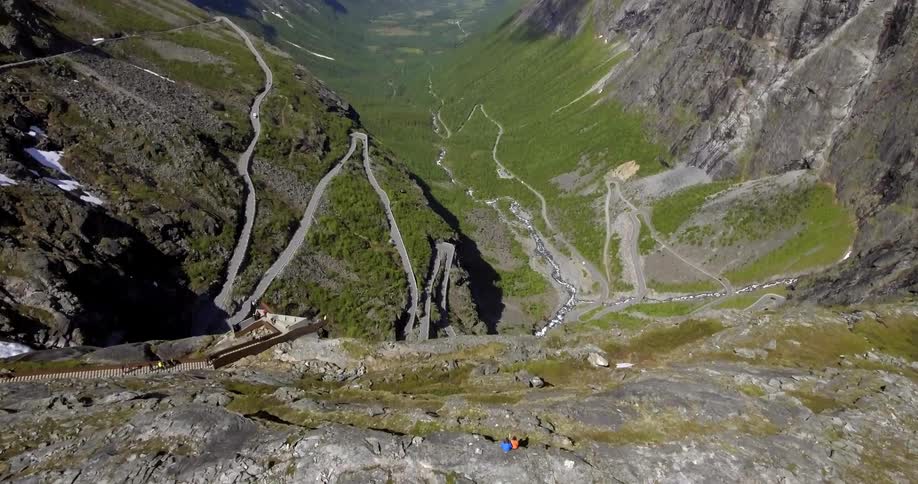 Two people sit on the edge of a tall cliff overlooking a winding road