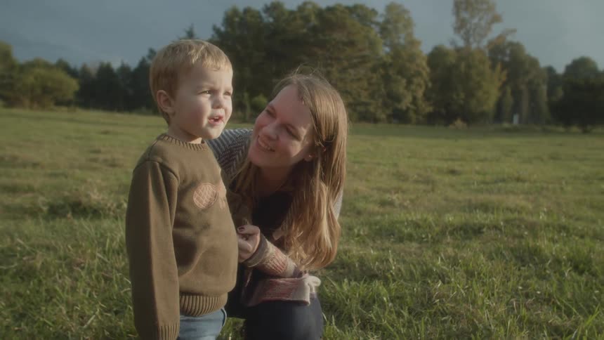 A mother in a field knees down and smiles at her son