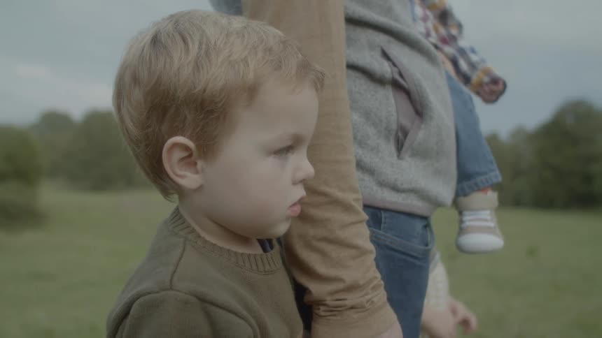 A woman stands outside on a field holding a baby in her arms and holding her young sons hand